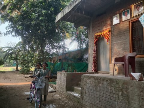 Woman with bicycle standing outside a brick rural house with green plants and vines decorating the entrance