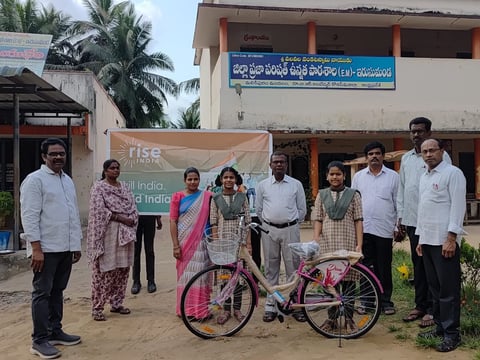 Group of people posing with a decorated bicycle in front of a building with a RISE India banner